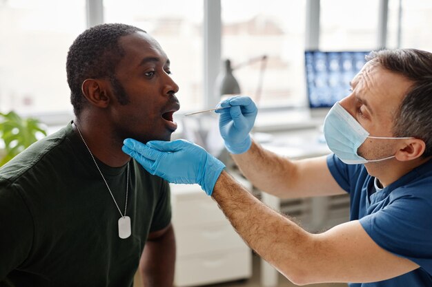 Junger schwarzer Militäroffizier, der mit weit geöffnetem Mund sitzt und einen PCR-Tupfertest auf Coronavirus in mod durchführt