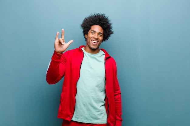 Junger schwarzer Sportmann, der glücklich, Spaß, überzeugt, positiv und rebellisch sich fühlt und Felsen oder Schwermetallzeichen mit der Hand gegen Schmutzwand macht