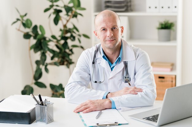Junger selbstbewusster Kliniker im weißen Kittel, der am Schreibtisch sitzt, während er Online-Daten über seine Patienten durchschaut