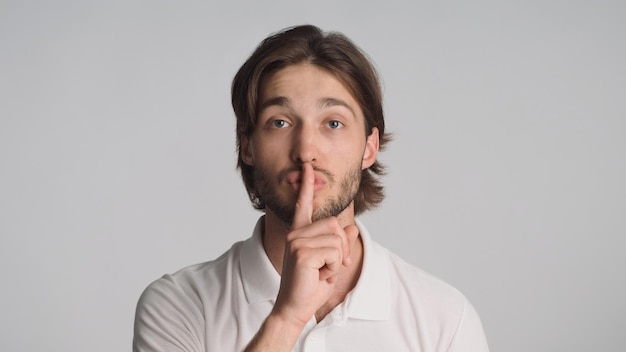 Junger selbstbewusster Mann, der den Zeigefinger auf den Lippen hält und darum bittet, vor der Kamera auf grauem Hintergrund zu schweigen Shh-Ausdruck Attraktiver Mann, der im Studio ein Schweigezeichen zeigt