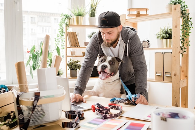 Junger seriöser Designer in Freizeitkleidung, der sich vor dem Laptop über den Tisch beugt, während er mit seinem Haustier im Internet nach neuen Ideen sucht