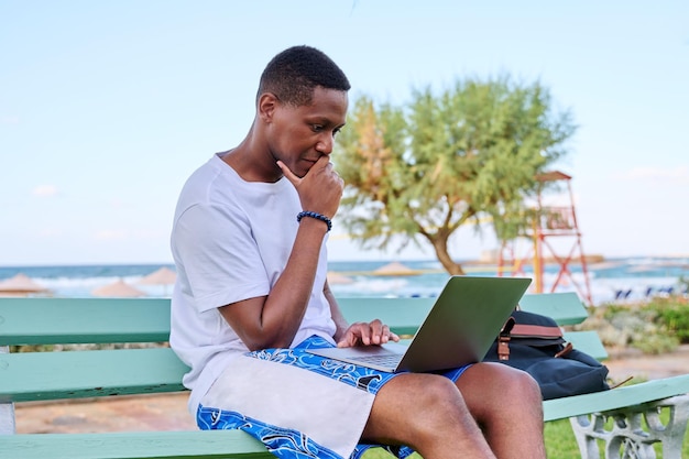 Junger seriöser männlicher Freiberufler, der sich am Strand entspannt und mit Laptop auf einer Bank sitzt