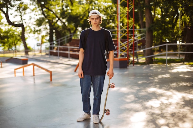 Junger Skater mit weißer Mütze schaut verträumt in die Kamera und hält Skateboard in der Hand mit großem, modernem Skatepark im Hintergrund