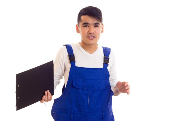 Junger, smart aussehender Mann mit dunklem Haar in weißem Hemd und blauem Overall mit weißem Stift und schwarzem Ordner und Gespräch auf weißem Hintergrund im Studio