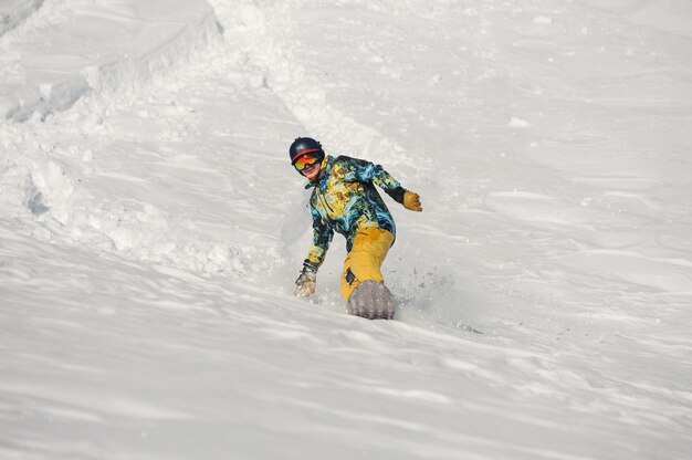 Junger Snowboarder in der hellen Sportbekleidung, die einen Schneehügel am hellen Wintertag hinunter reitet