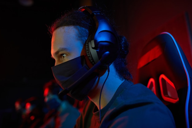 Junger Spieler in Kopfhörern und in Schutzmaske, die Computerspiele im dunklen Raum spielen