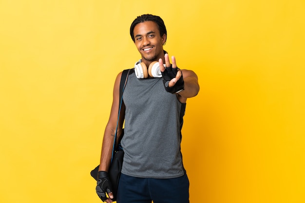 Junger Sport Afroamerikaner Mann mit Zöpfen mit Tasche lokalisiert auf gelbem Hintergrund glücklich und zählt drei mit den Fingern