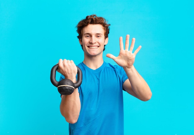 Junger Sportjunge, der freundlich lächelt und aussieht, Nummer fünf oder fünf mit der Hand nach vorne zeigt, herunterzählt. Hantelkonzept