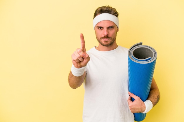 Junger sportkaukasier, der zu yogakursen geht, während er eine matte hält, die auf gelbem hintergrund isoliert ist und die nummer eins mit dem finger zeigt.