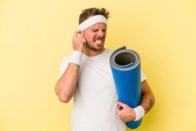 Junger sportkaukasier, der zu yogakursen geht, während er eine matte hält, die auf gelbem hintergrund isoliert ist und die ohren mit den händen bedeckt.