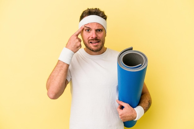 Junger sportkaukasier, der zu yogakursen geht, während er eine matte isoliert auf gelbem hintergrund hält, die eine enttäuschungsgeste mit dem zeigefinger zeigt