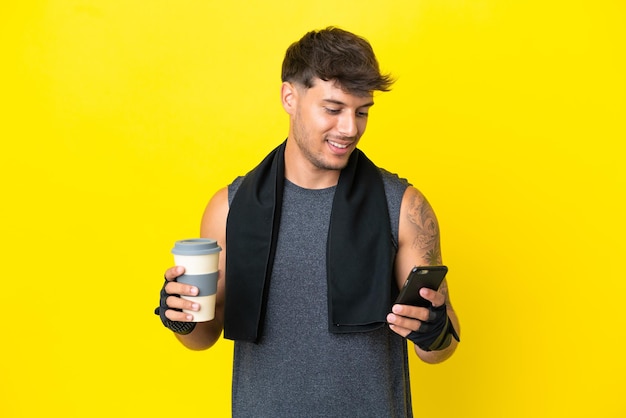 Junger sportkaukasischer Mann mit Handtuch isoliert auf gelbem Hintergrund, der Kaffee zum Mitnehmen und ein Handy hält