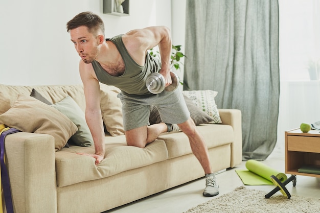 Junger Sportler in Aktivkleidung, der sich auf Couch stützt, während Übung mit Hanteln für Arme und Bizeps während des morgendlichen Trainings zu Hause macht