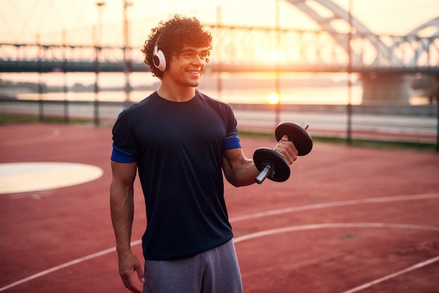 Junger sportlicher fit Mann, der Gewicht in seinem Arm hält, während er die Musik hört. Außerhalb des Trainings am frühen Morgen.