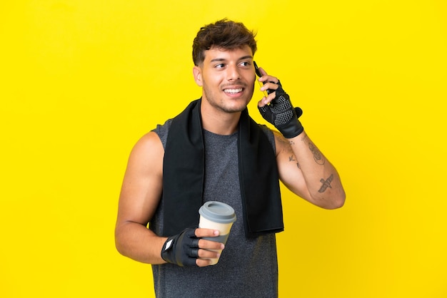 Junger sportlicher kaukasischer Mann mit Handtuch isoliert auf gelbem Hintergrund, der Kaffee zum Mitnehmen und ein Handy hält