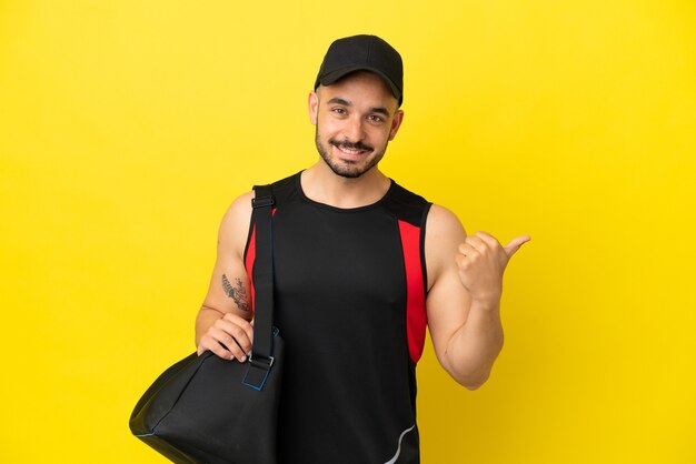 Junger sportlicher kaukasischer Mann mit Sporttasche isoliert auf gelbem Hintergrund, der auf die Seite zeigt, um ein Produkt zu präsentieren
