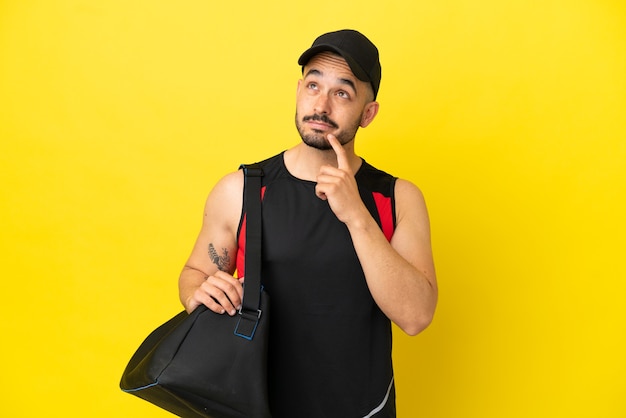 Junger sportlicher kaukasischer Mann mit Sporttasche isoliert auf gelbem Hintergrund, der beim Nachschlagen Zweifel hat