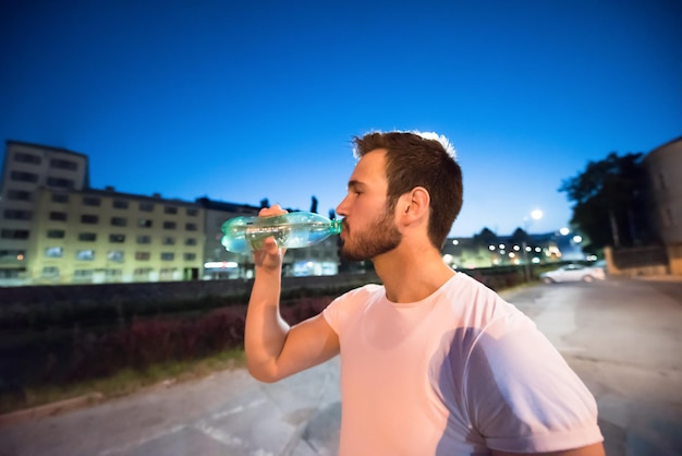 Junger sportlicher Mann, der nach einer nächtlichen Laufeinheit in der Stadt Wasser trinkt