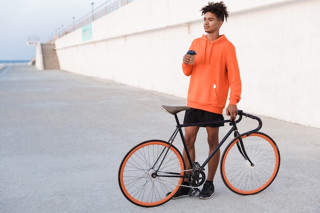 Junger Sportmann draußen am Strand mit Fahrrad, das Kaffee hält