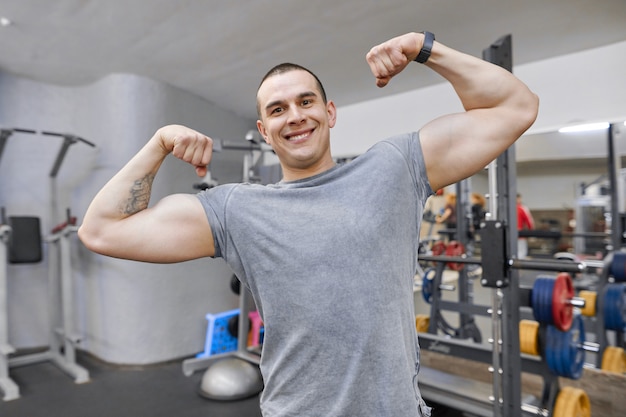Junger starker lächelnder muskulöser Mann in der Turnhalle, die starke muskulöse Arme zeigt.