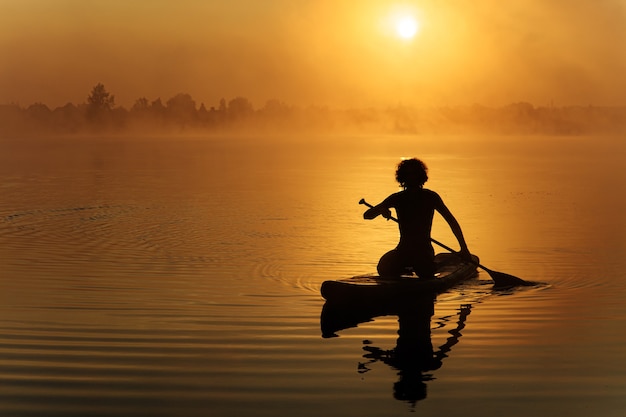 Junger starker Mann in der Silhouette, die Paddel für das Schwimmen auf sup Brett am nebligen See verwendet.