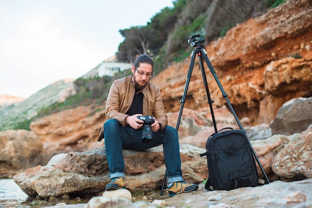 Junger stilvoller Fotograf, der auf Felsen sitzt und Fotos aufpasst, die er auf seiner Kamera machte