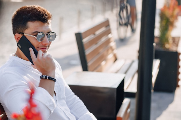 Junger stilvoller Kerl im Hemd mit Telefon auf Bank am sonnigen warmen Tag draußen