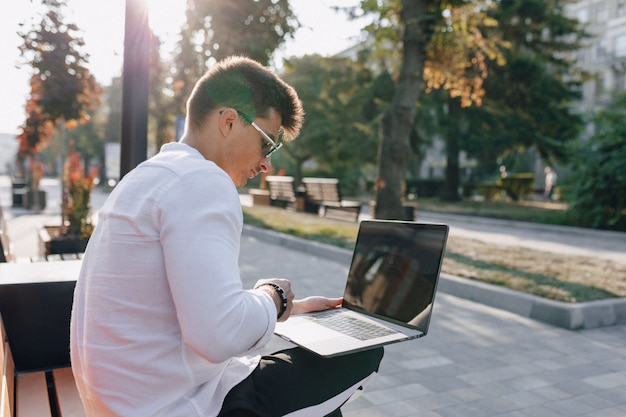Junger stilvoller Kerl im Hemd mit Telefon und Notizbuch auf Bank am sonnigen warmen Tag im Freien, freiberuflich