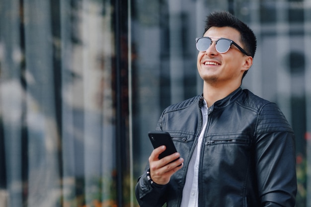 Junger stilvoller Kerl in der Brille in der schwarzen Lederjacke mit Telefon auf Glas