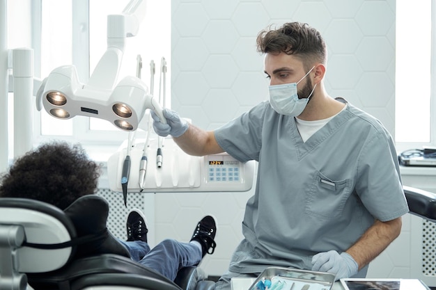 Junger Stomatologe in Arbeitskleidung sitzt neben kleinem Patienten