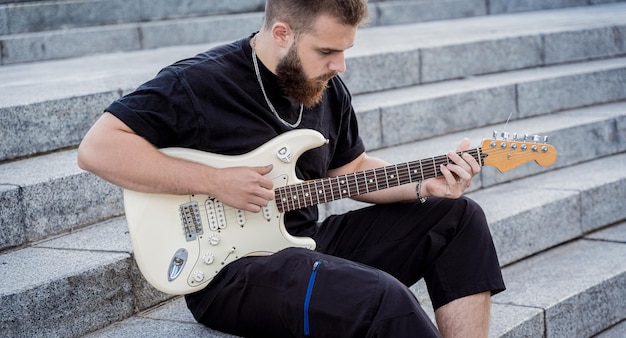 Junger Straßenmusiker, der Gitarre spielt, sitzt auf Granitstufen