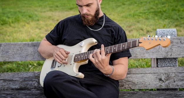 Junger Straßenmusiker, der Gitarre spielt, während er auf der Bank sitzt