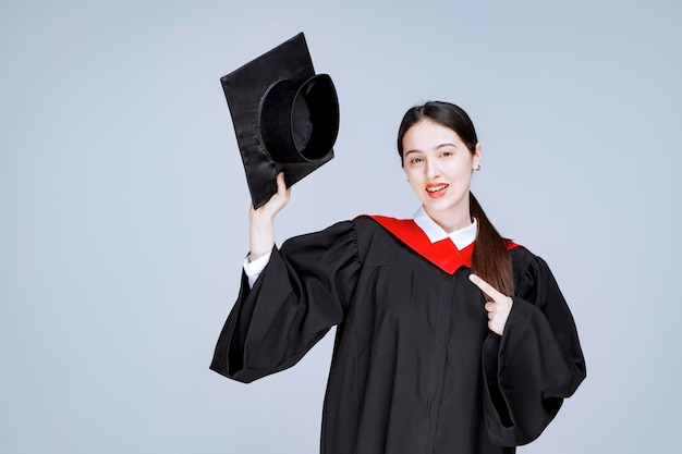 Junger Student im Kleid, das auf Kappe zeigt. Foto in hoher Qualität