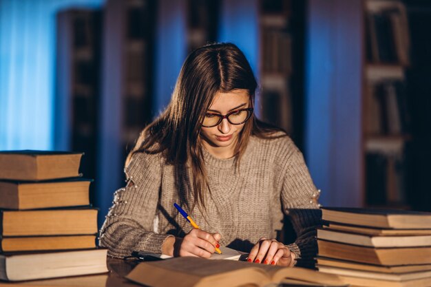 Junger Student in Gläsern, die sich auf die Prüfung vorbereiten. Mädchen am Abend sitzt an einem Tisch in der Bibliothek mit einem Stapel Bücher