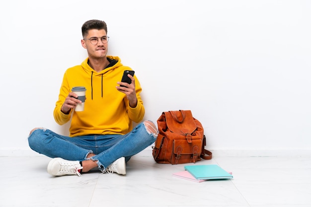 Junger Student kaukasischer Mann sitzt auf dem Boden isoliert auf weißem Hintergrund und hält Kaffee zum Mitnehmen und ein Handy, während er etwas denkt