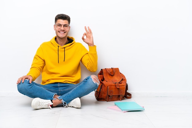 Junger Student kaukasischer Mann sitzt auf dem Boden isoliert auf weißem Hintergrund und zeigt mit den Fingern ein Ok-Zeichen