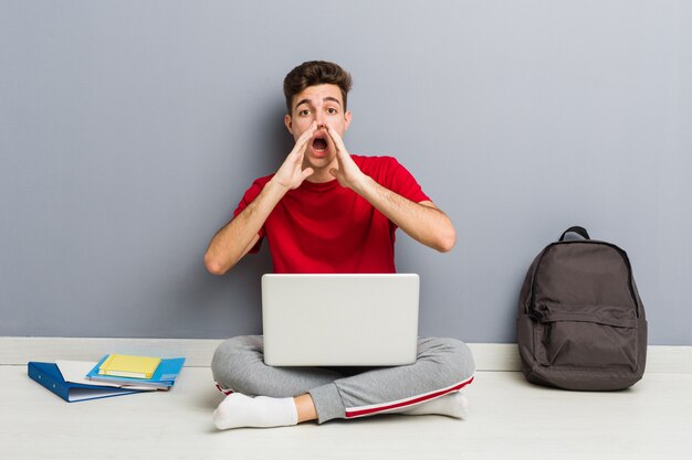 Junger Studentenmann, der auf seinem Hausboden hält einen Laptop sitzt