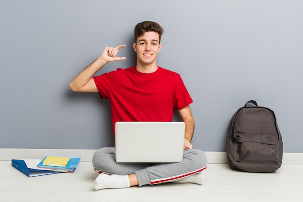 Junger studentenmann, der auf seinem hausboden hält einen laptop sitzt