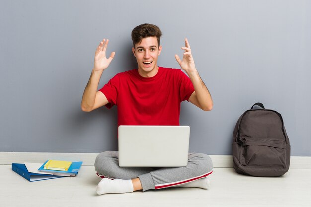 Junger Studentenmann, der auf seinem Hausboden hält einen Laptop sitzt