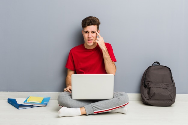 Junger Studentenmann, der auf seinem Hausboden hält einen Laptop sitzt