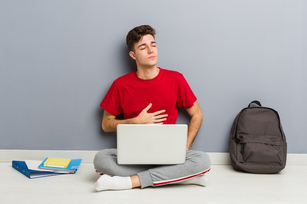 Junger Studentenmann, der auf seinem Hausboden hält einen Laptop sitzt