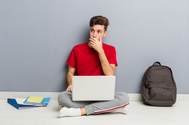 Junger Studentenmann, der auf seinem Hausboden hält einen Laptop sitzt