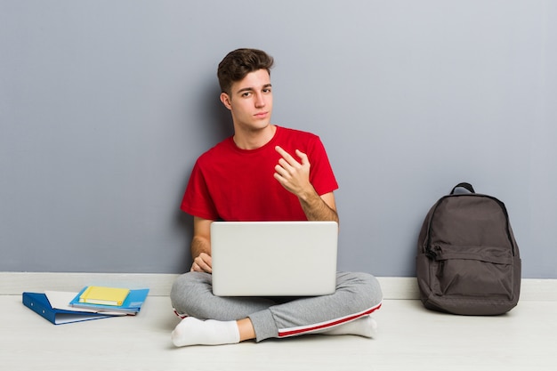 Junger Studentenmann, der auf seinem Hausboden hält einen Laptop sitzt