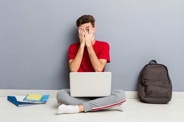 Junger Studentenmann, der auf seinem Hausboden hält einen Laptop sitzt