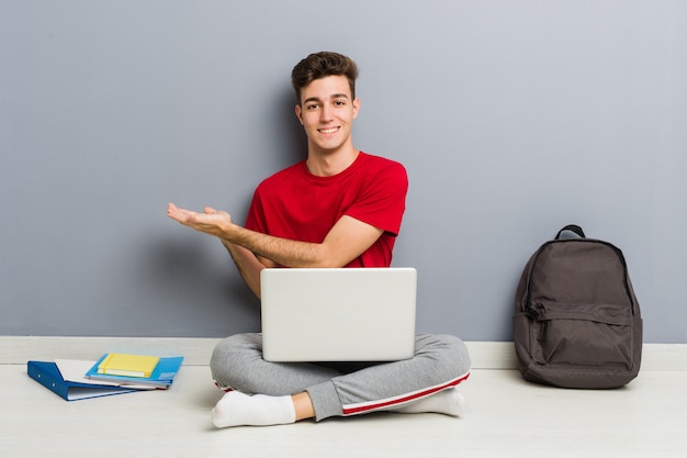 Junger Studentenmann, der auf seinem Hausboden hält einen Laptop sitzt