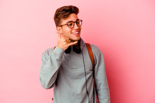 Junger Studentenmann einzeln auf rosafarbenem Hintergrund, der eine Handyanrufgeste mit den Fingern zeigt.