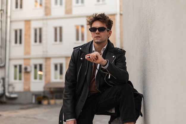 Junger stylischer Hipster in trendiger Kleidung in Vintage-Sonnenbrillen ruht sich aus und genießt Nikotin in der Nähe der Wand auf der Straße. Cooler amerikanischer Kerl raucht draußen eine Zigarette. Entspannende, aber schädliche Gewohnheit.