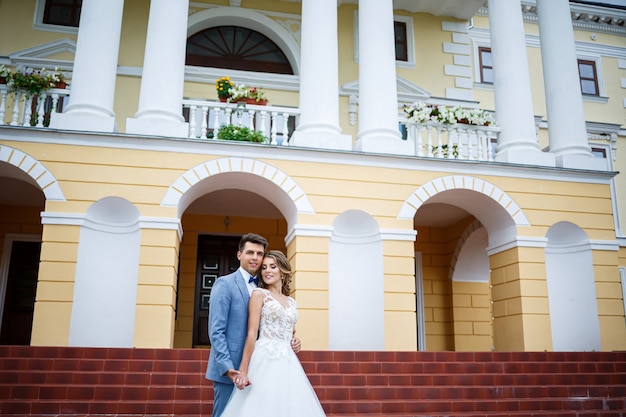 Junger stylischer Kerl im Kostüm des Bräutigams und der Braut schönes Mädchen in einem weißen Kleid mit einem Zug, der auf dem Hintergrund eines großen Hauses mit Säulen an ihrem Hochzeitstag spaziert