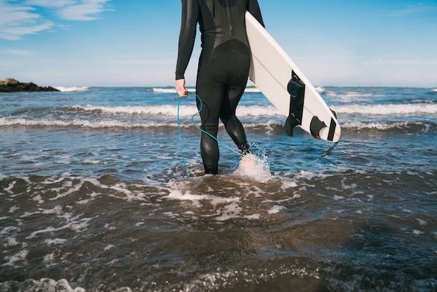 Junger Surfer, der mit seinem Surfbrett in einem schwarzen Surfanzug ins Wasser tritt. Sport- und Wassersportkonzept.