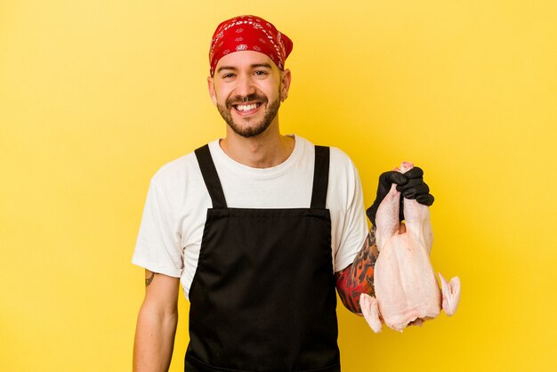 Junger tätowierter kaukasischer batcher-mann, der ein huhn lokalisiert auf gelbem hintergrund glücklich, lächelnd und fröhlich hält.